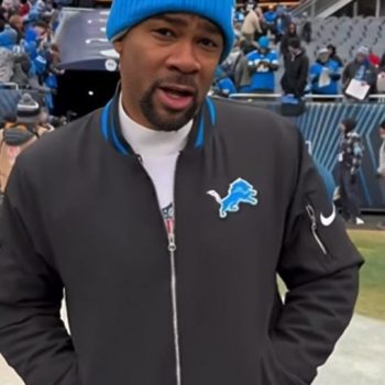 Person wearing a black jacket with a logo, blue beanie, and white shirt at a sports event.