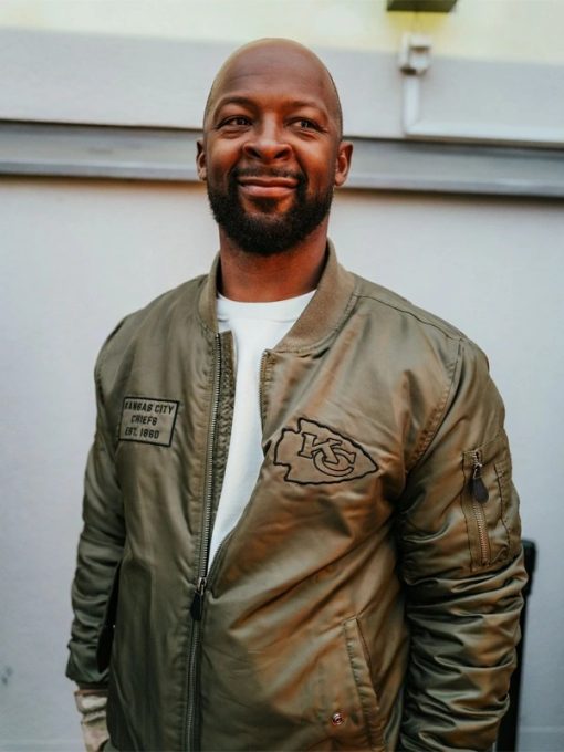 Dante-Hall-Chiefs-Military-Bomber-Jacket-510x680-1 Man wearing a green bomber jacket with a logo, standing against a light-colored wall in UK